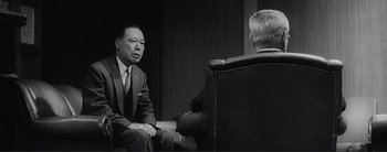 Movie still from “The Bad Sleep Well” (1960), directed by Akira Kurosawa – A man in a suit and tie sitting in front of another man in a chair; Medium shot, Over the shoulder angle