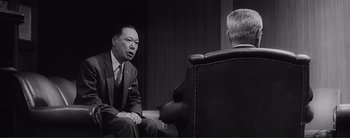 Movie still from “The Bad Sleep Well” (1960), directed by Akira Kurosawa – A man in a suit and tie sitting next to another man in a chair; Medium shot, Over the shoulder angle