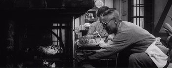 Movie still from “The Bad Sleep Well” (1960), directed by Akira Kurosawa – A man sitting in front of a fireplace while eating; Medium shot, Over the shoulder angle