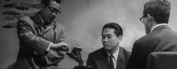 Movie still from “The Bad Sleep Well” (1960), directed by Akira Kurosawa – A man is looking at a cell phone while another man looks on; Medium shot, Low angle