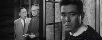 Movie still from “The Bad Sleep Well” (1960), directed by Akira Kurosawa – A man standing next to another man in front of a window; Close Up shot, Low angle