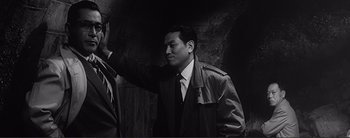 Movie still from “The Bad Sleep Well” (1960), directed by Akira Kurosawa – A black and white photo of a man in a trench coat; Medium shot, Over the shoulder angle