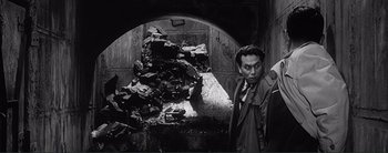 Movie still from “The Bad Sleep Well” (1960), directed by Akira Kurosawa – A black and white photo of a man standing in front of a pile of rubble; Medium shot, High angle