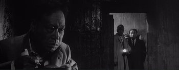 Movie still from “The Bad Sleep Well” (1960), directed by Akira Kurosawa – A man standing in front of a door next to another man holding a flashlight; Close Up shot, Low angle