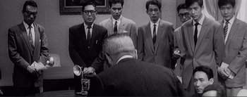Movie still from “The Bad Sleep Well” (1960), directed by Akira Kurosawa – A group of men in suits and ties standing in front of a man; Medium shot, Low angle