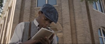 Movie still from “The Banker” (2020), directed by George Nolfi – A young man reading a book in front of a brick building; Close Up shot, Low angle