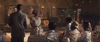 Movie still from “The Banker” (2020), directed by George Nolfi – A woman in a white dress standing in front of a group of people; Wide shot, High angle