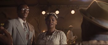 Movie still from “The Banker” (2020), directed by George Nolfi – A woman wearing a floral head wreath standing in front of a group of people; Medium shot, Low angle