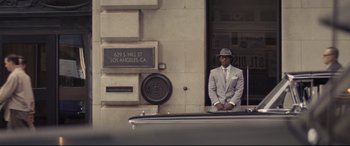 Movie still from “The Banker” (2020), directed by George Nolfi – A man in a suit and hat standing in front of a building; Wide shot, Low angle