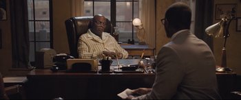 Movie still from “The Banker” (2020), directed by George Nolfi – A man sitting at a desk in front of another man; Medium shot, Over the shoulder angle