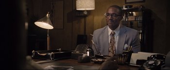 Movie still from “The Banker” (2020), directed by George Nolfi – A man sitting at a table in front of a lamp; Medium shot, Over the shoulder angle
