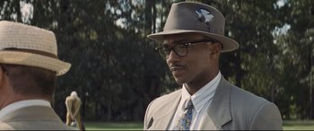 Movie still from “The Banker” (2020), directed by George Nolfi – A man in a suit and hat is standing in a park; Close Up shot, Over the shoulder angle