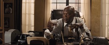 Movie still from “The Banker” (2020), directed by George Nolfi – A man sitting at a desk holding a glass; Medium shot, High angle