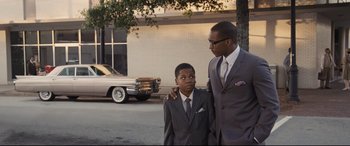 Movie still from “The Banker” (2020), directed by George Nolfi – A man and a young boy in suits standing next to each other in front of an old car; Medium shot, Over the shoulder angle