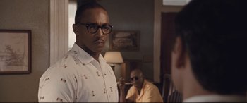 Movie still from “The Banker” (2020), directed by George Nolfi – A man with glasses standing next to another man in a living room; Medium shot, Over the shoulder angle