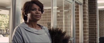 Movie still from “The Banker” (2020), directed by George Nolfi – A woman standing in front of a building holding a feather duster; Close Up shot, Low angle