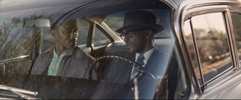Movie still from “The Banker” (2020), directed by George Nolfi – A man in a suit and hat sitting in the driver's seat of a car; Medium shot, Over the shoulder angle