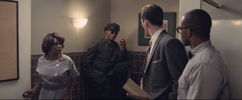Movie still from “The Banker” (2020), directed by George Nolfi – A man in a suit and a tie in a bathroom; Medium shot, Low angle