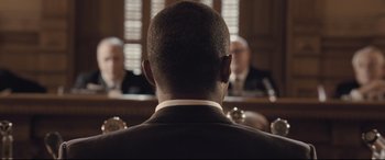 Movie still from “The Banker” (2020), directed by George Nolfi – A man in a suit and tie in front of a group of people; Close Up shot, Over the shoulder angle