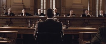 Movie still from “The Banker” (2020), directed by George Nolfi – A man sitting in front of a group of people in a courtroom; Wide shot, Over the shoulder angle
