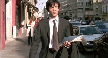 Movie still from “The Beat That My Heart Skipped” (2005), directed by Jacques Audiard – A man in a suit and tie holding a book walking down the street; Medium shot, Low angle