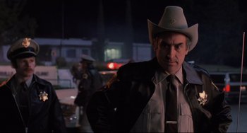 Movie still from “The Blob” (1988), directed by Chuck Russell – A man in a cowboy hat standing in front of a police car; Medium shot, Low angle