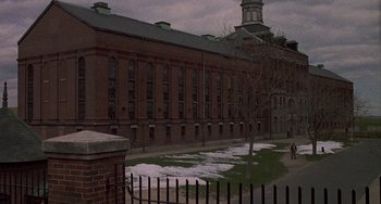 Movie still from “The Brink's Job” (1978), directed by William Friedkin – A brick building with snow on the ground and a fence; Extreme Wide shot, High angle