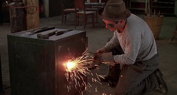 Movie still from “The Brink's Job” (1978), directed by William Friedkin – A man wearing a hat and glasses is using a grinder to make sparks; Medium shot, Low angle