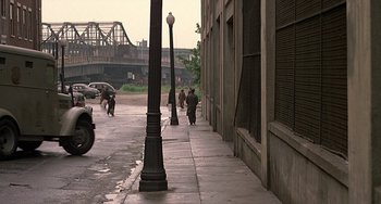 Movie still from “The Brink's Job” (1978), directed by William Friedkin – People are walking down the sidewalk on a rainy day; Extreme Wide shot, Over the shoulder angle