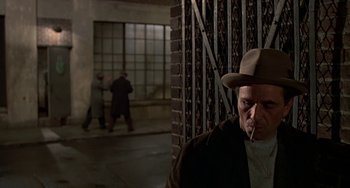 Movie still from “The Brink's Job” (1978), directed by William Friedkin – A man in a hat and a brown jacket smoking a cigarette; Medium shot, Over the shoulder angle