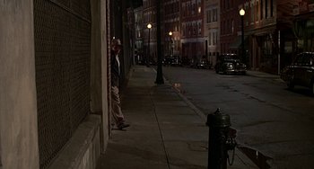 Movie still from “The Brink's Job” (1978), directed by William Friedkin – A man leaning against a wall on a sidewalk at night; Wide shot, Over the shoulder angle