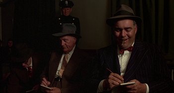 Movie still from “The Brink's Job” (1978), directed by William Friedkin – A man in a suit and hat writing on a paper; Medium shot, Low angle