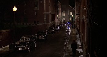 Movie still from “The Brink's Job” (1978), directed by William Friedkin – A man standing on the side of a road next to a row of parked cars; Extreme Wide shot, High angle