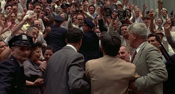 Movie still from “The Brink's Job” (1978), directed by William Friedkin – A group of people standing around each other in front of a crowd of onlookers; Wide shot, High angle