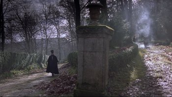 Movie still from “The Duellists” (1977), directed by Ridley Scott – A man in a long black coat walking in the woods near a pillar; Wide shot, High angle