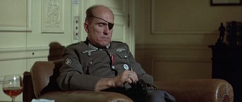 Movie still from “The Eagle Has Landed” (1976), directed by John Sturges – A man in uniform sitting on a couch looking at his watch; Medium shot, High angle