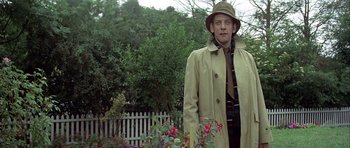 Movie still from “The Eagle Has Landed” (1976), directed by John Sturges – A man wearing a hat and a trench coat standing next to flowers; Medium shot, Low angle