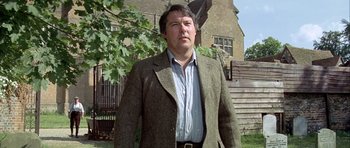 Movie still from “The Eagle Has Landed” (1976), directed by John Sturges – A man in a suit and tie standing in front of a building; Medium shot, Low angle