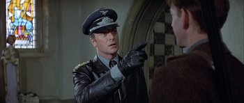 Movie still from “The Eagle Has Landed” (1976), directed by John Sturges – A man in a leather jacket and a hat points to someone; Close Up shot, Over the shoulder angle