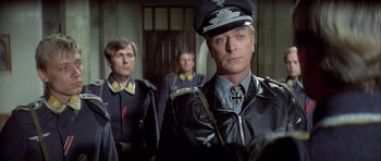 Movie still from “The Eagle Has Landed” (1976), directed by John Sturges – A man in a military uniform stands in front of two other men; Medium shot, Over the shoulder angle