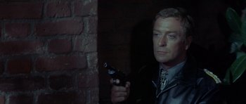 Movie still from “The Eagle Has Landed” (1976), directed by John Sturges – A man holding a gun while standing next to a brick wall; Close Up shot, Over the shoulder angle