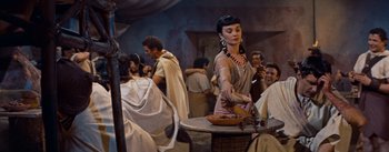 Movie still from “The Egyptian” (1954), directed by Michael Curtiz – A woman is holding a bowl of food in front of a group of people; Medium shot, Over the shoulder angle