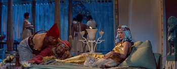 Movie still from “The Egyptian” (1954), directed by Michael Curtiz – A couple of people that are sitting on a bed with a cat; Wide shot, High angle