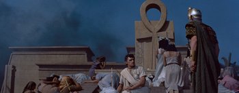 Movie still from “The Egyptian” (1954), directed by Michael Curtiz – A man sitting on steps next to a statue of an anunnaki god; Wide shot, Low angle