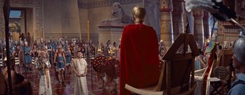 Movie still from “The Egyptian” (1954), directed by Michael Curtiz – A man wearing a red robe and a red cape is sitting in front of a group of people; Extreme Wide shot, High angle