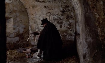 Movie still from “The Enigma of Kaspar Hauser” (1974), directed by Werner Herzog – A man in a black coat and a black hat sitting on the ground; Wide shot, High angle
