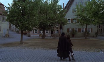 Movie still from “The Enigma of Kaspar Hauser” (1974), directed by Werner Herzog – Two people walking down the sidewalk in front of a house; Wide shot, Over the shoulder angle
