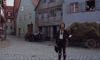 Movie still from “The Enigma of Kaspar Hauser” (1974), directed by Werner Herzog – A man standing on a cobblestone street in front of a horse drawn carriage; Wide shot, Low angle