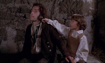 Movie still from “The Enigma of Kaspar Hauser” (1974), directed by Werner Herzog – A man and a boy are sitting in front of a stone wall; Medium shot, Over the shoulder angle