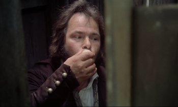 Movie still from “The Enigma of Kaspar Hauser” (1974), directed by Werner Herzog – A man with long black hair and a beard is holding his hand to his mouth; Medium shot, Low angle